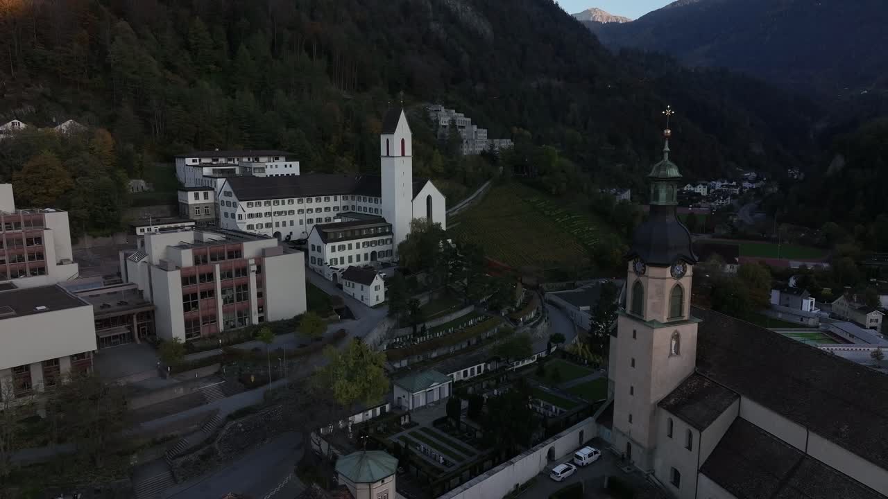 Drone flies towards and passes near church in Chur town during morning with majestic mountains in background, Graubünden, Switzerland.