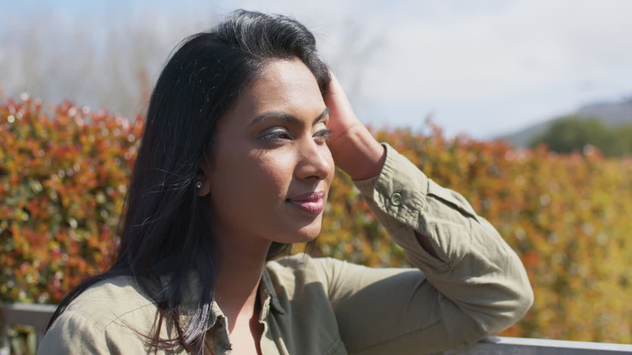 Breeze ruffling hair, mid adult Indian woman tucking hair, smiling on bench by autumn hedge
