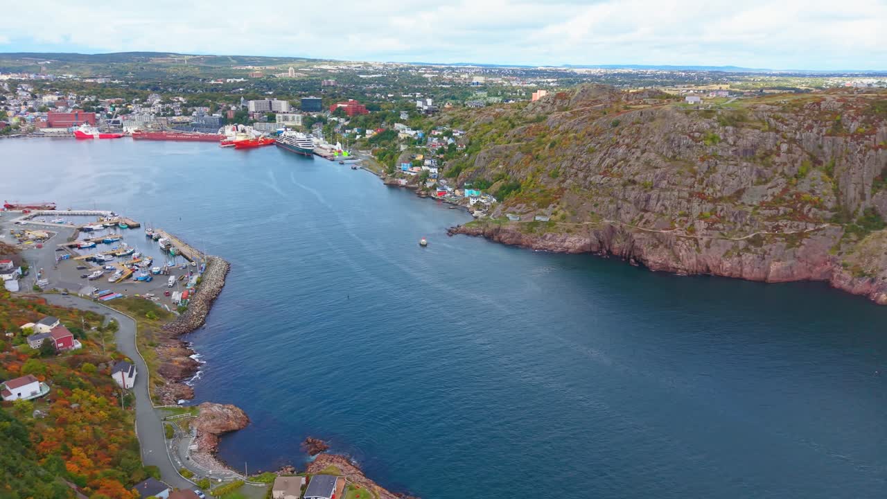 Drone view of Fort Amherst’s calm bay in autumn reveals teal water, rocky cliffs, cosy hillside homes and vibrant foliage. The endless horizon frames this rugged coastline with maritime charm