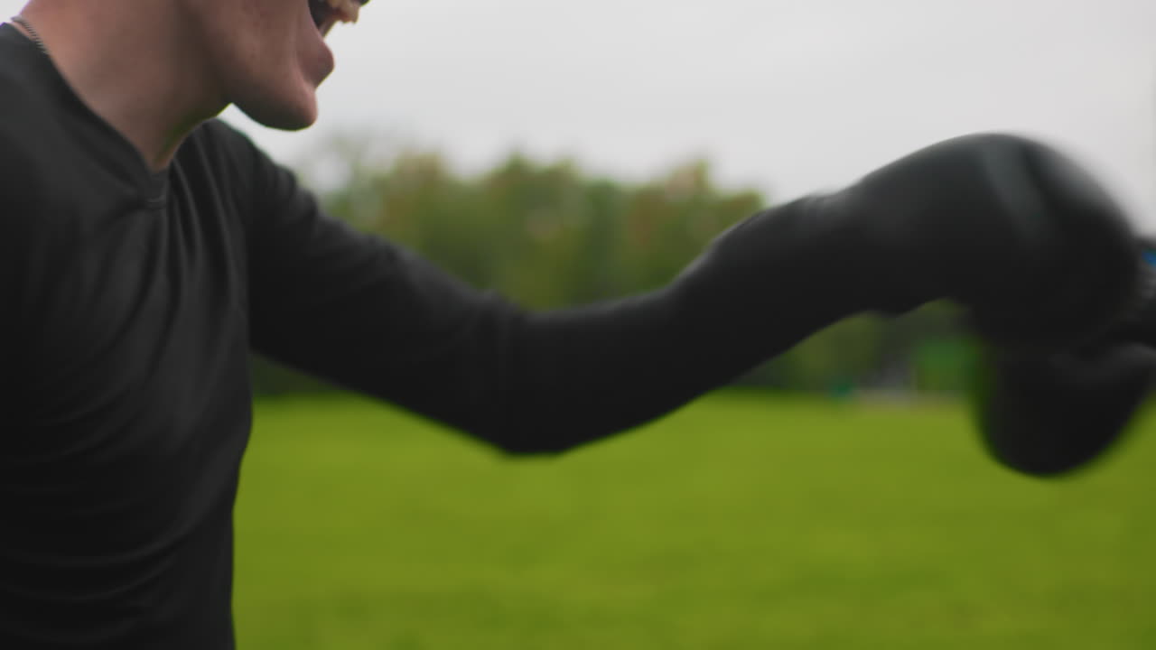 outdoor boxing sparring gloves green field young man in black top wearing gloves throws controlled punches at trainer mitts closeup midshot overcast sky lush background rhythmic motion concentrated