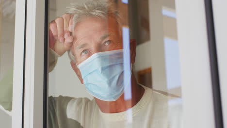 Hombre-Caucásico-Mayor-Con-Mascarilla-Mirando-Por-La-Ventana-En-Casa