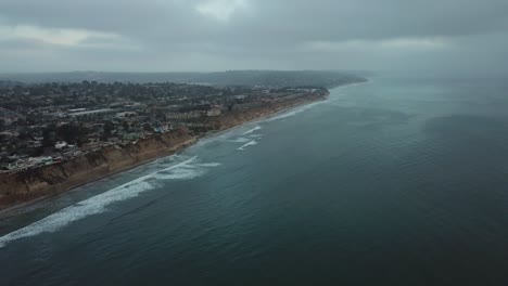 Vista-Desde-Un-Dron-Que-Se-Inclina-Hacia-Abajo-Sobre-El-Océano-Mostrando-El-Mar,-La-Playa-Y-Un-Pueblo-Cerca-Del-Borde-De-Un-Acantilado