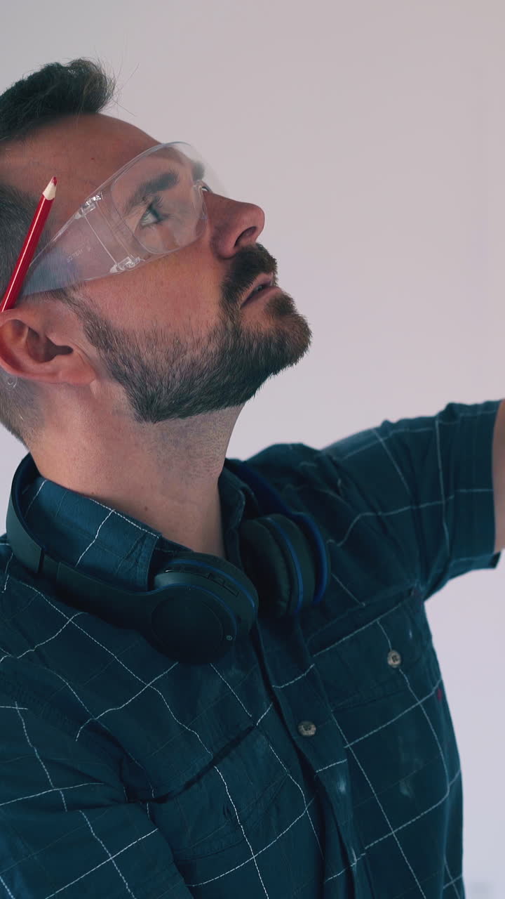 Dolly cam: young bearded handyman in goggle checks white wall angle using level and pencil at renewal light empty room