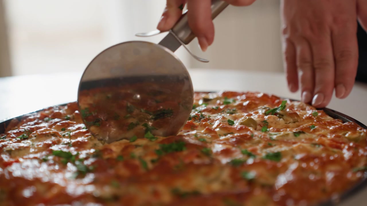 Vista ravvicinata di una mano che preme una rotella tagliapizza in metallo attraverso formaggio fuso e crosta dorata su una teglia rotonda, con i condimenti che si spostano leggermente ad ogni fetta, in una calda luce da cucina interna.