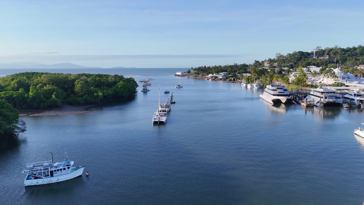 Drone footage captures boats in a serene marina at Port Douglas, Australia, under clear skies with lush greenery and calm waters