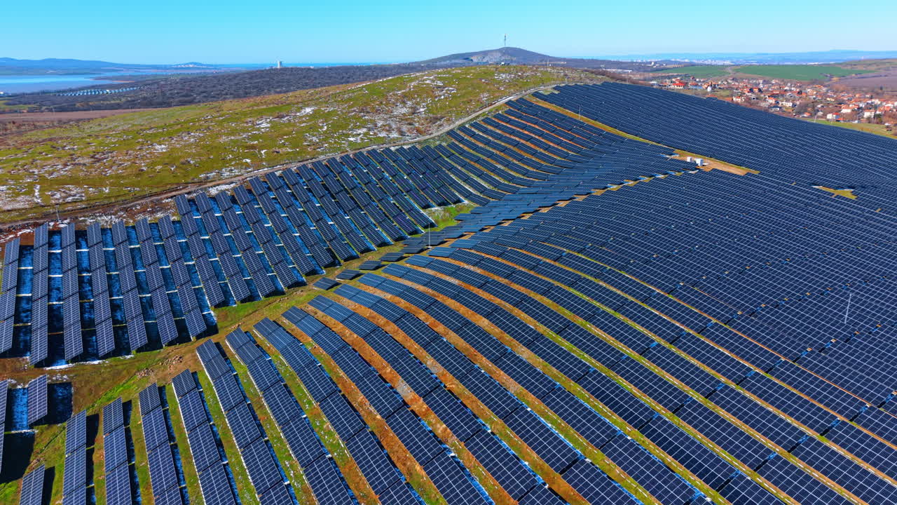 Massivt solenergianlæg i landskab nær sø. Bredt luftperspektiv af solpaneler, der dækker bakkeskråninger, med en fjern sø og horisont