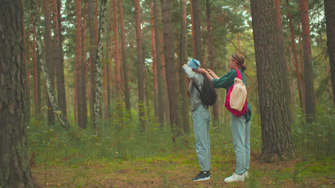 dos mujeres jóvenes en una escena de bosque tranquilo, una vestida con una camisa azul con una bolsa roja ayuda a una amiga con un pañuelo azul a ponerse su bolsa negra, rodeada de árboles altos y vegetación exuberante