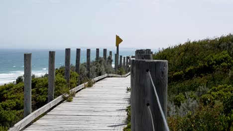 Camino-De-Madera-Para-Caminar-Y-Caminar-Cerca-De-La-Playa,-El-Viento-Sopla-Sobre-La-Hierba-En-El-Fondo