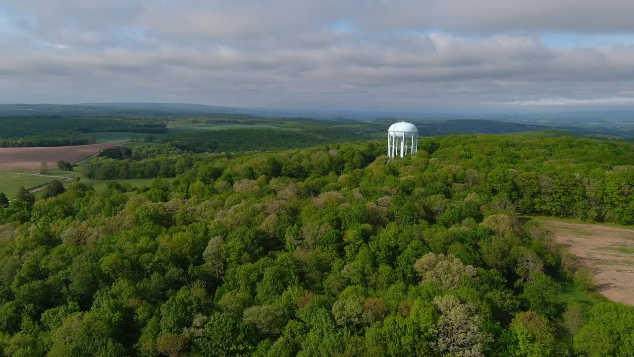 Vista aerea dall'alto con drone di una torre dell'acqua bianca che si erge sopra una fitta foresta nel Parco Statale di Mount Pisgah, circondata da dolci colline e terreni agricoli sotto un cielo nuvoloso.