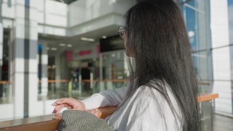 rear view of woman with long black hair resting on wooden rail while gently tapping her finger, wearing white shirt and glasses, in sunlit modern shopping mall with elegant interior and relaxed mood