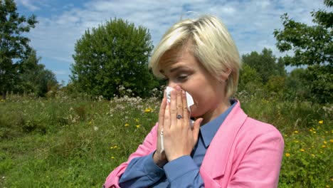 mujer con fiebre del heno estornudando y soplando en un pañuelo mientras estaba en un prado verde con flores, usando un cárdigan rosa, camisa azul, cabello rubio, maquillaje claro y ojos verdes