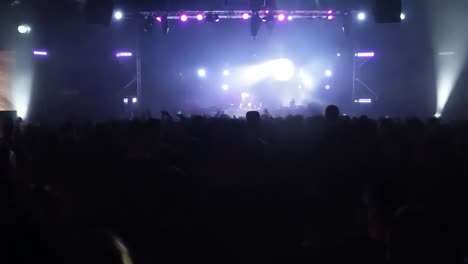 una gran multitud de personas en un concierto con una banda en el escenario bajo luces brillantes