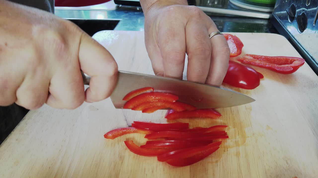 A man finely slices a red bell pepper on a wooden chopping board in a home kitchen whilst preparing dinner