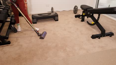 a woman vacuums the carpet in a home gym, surrounded by fitness equipment. bright lighting enhances the clean, organized space