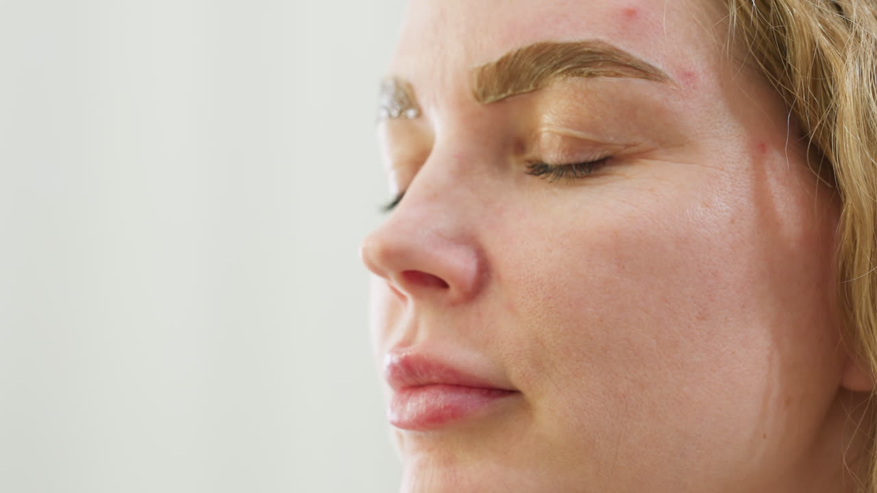 Beautician in black gloves uses cotton swab to gently clean excess cream beneath client eyebrow during beauty procedure. Close-up highlights precision and care in modern cosmetic treatment session