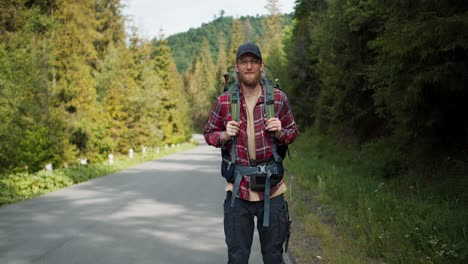 A-tourist-in-special-clothing-for-hiking-in-a-red-shirt-stands-against-the-backdrop-of-a-green-forest.-Male-traveler-blond-with-beard-wearing-glasses