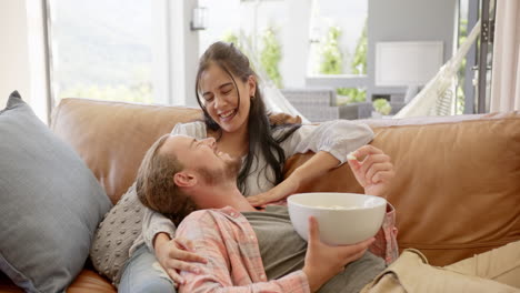 A-young-biracial-couple-shares-a-moment-of-laughter-on-a-sofa-at-home,-eating-popcorn