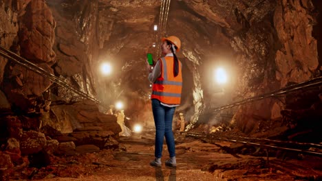full body back view of asian female engineer with safety helmet working on a green screen smartphone and looking around in underground mine tunnel