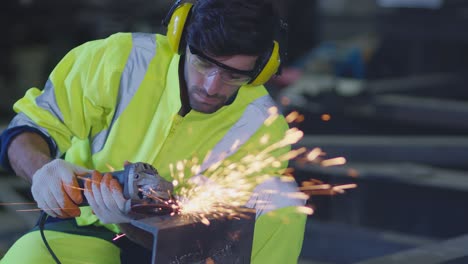 trabajador masculino caucásico en un uniforme seguro usar soldadores cueros, rueda eléctrica molienda en la estructura de acero en el factor en el garaje, chispas bokeh naranja vuelan a los lados