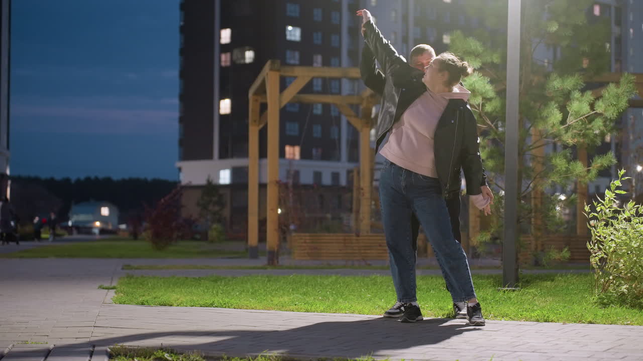 Husband and wife enjoying playful dance under street light, wind gently blowing trees around them, romantic and joyful moment outdoors in the evening