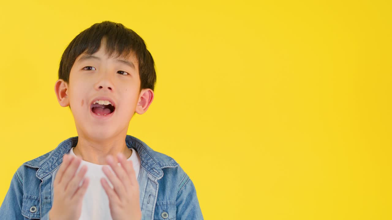 Young boy in denim jacket expresses surprise, hands on face, brightly lit yellow studio background
