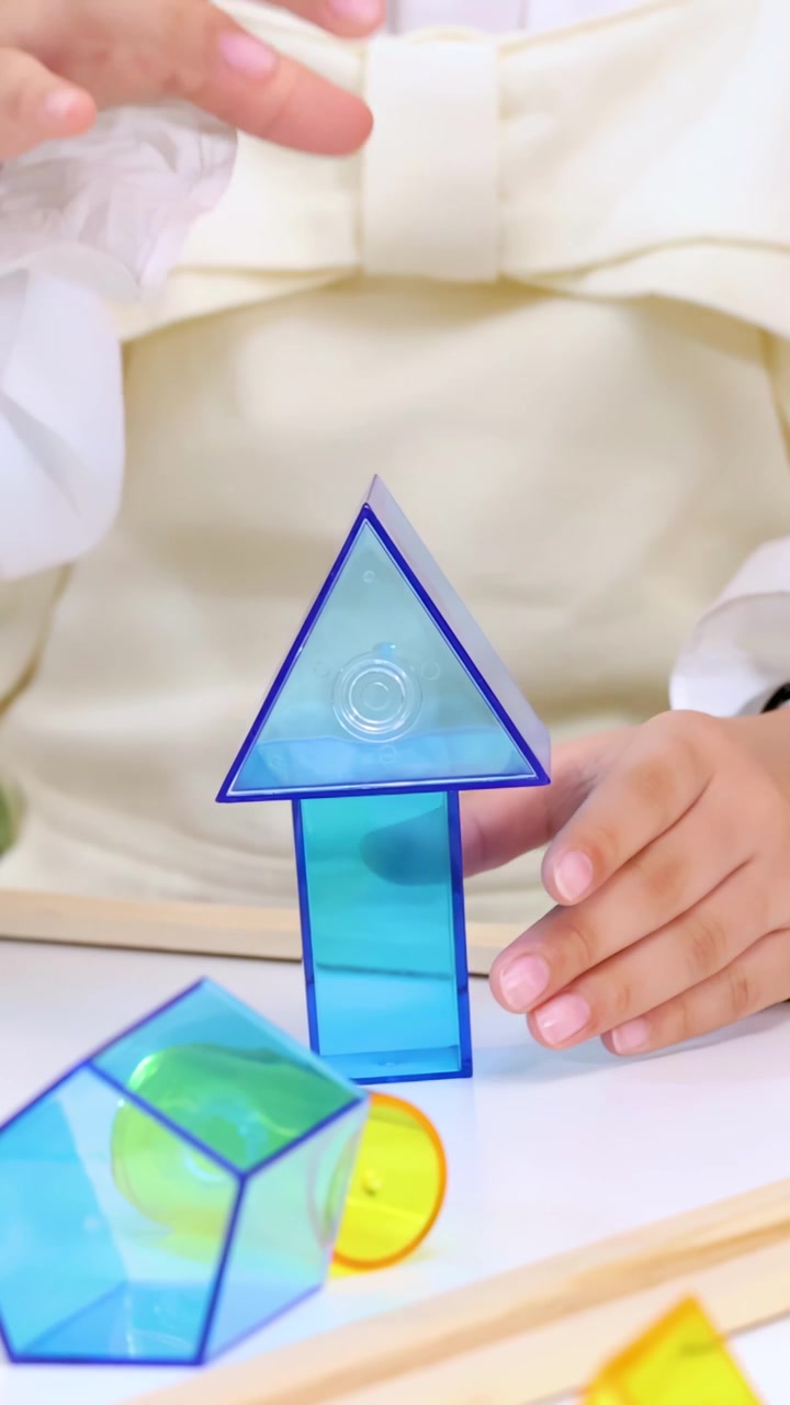 Child’s hands assemble transparent geometric blocks into tower structure on bright classroom table, static shot