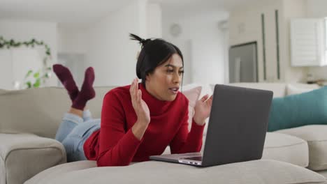 video de una feliz mujer biracial en hijab acostada en el sofá y usando una computadora portátil
