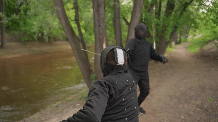 Due persone con equipaggiamento protettivo nero da scherma si fronteggiano su un sentiero sterrato circondato da alberi, preparandosi o impegnandosi in un combattimento con la spada. Una persona è visibile da dietro, con indosso un elmetto.
