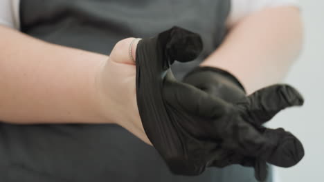 Close-up of beautician putting on black glove with silver ring on thumb, dressed in dark apron, preparing for work in clean salon space with blurred background and lamp glowing softly at side
