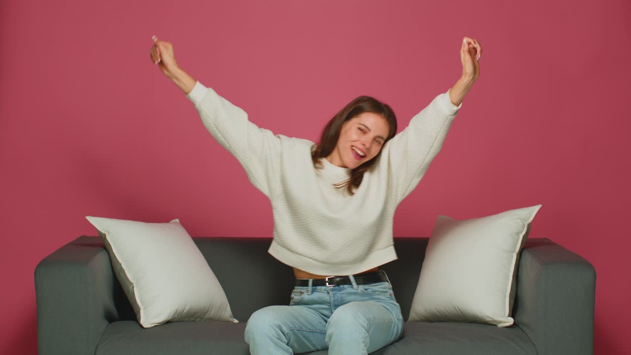 즐거운 젊은 소녀가 춤을 추고 음악을 듣고, 재미를 느끼고, 승리를 축하하며, 분홍색 스튜디오 배경에 소파에 앉아 있습니다.