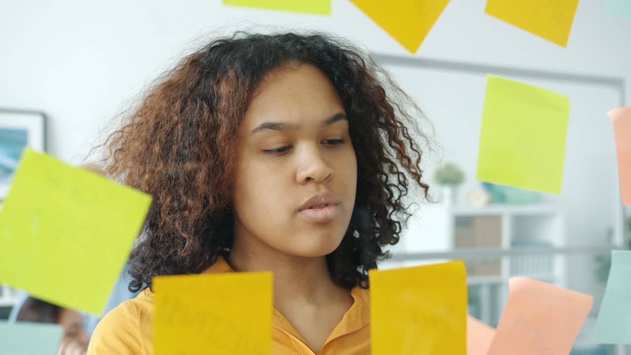 Close-up of serious Afro-American girl taking colorful sticky notes off glassboard in office