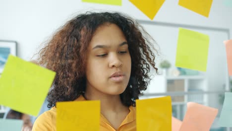 close-up of serious afro-american girl taking colorful sticky notes off glassboard in office