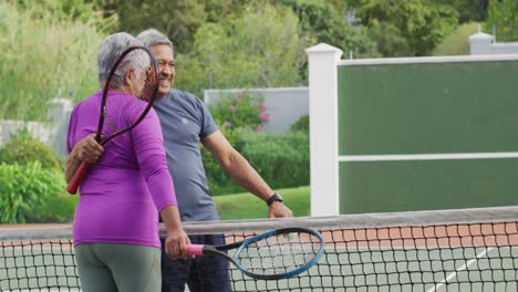video de una feliz pareja de alto nivel biracial sosteniendo raquetas y abrazándose en la cancha de tenis