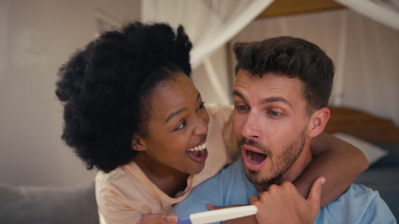 Close Up Of Excited Couple In Bedroom At Home Celebrating Positive Pregnancy Test Result