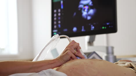 female doctor's hand moving the sensor by the big pregnant belly. black screen of a sonographer at backdrop in blur.