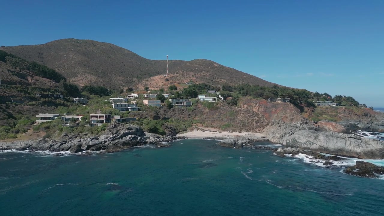 Voo avançando sobre uma enseada turquesa com costas rochosas revelando vilas exclusivas no topo do penhasco aninhadas em uma encosta de matagal seco sob um céu azul claro. Punta Pite, Papudo, Chile