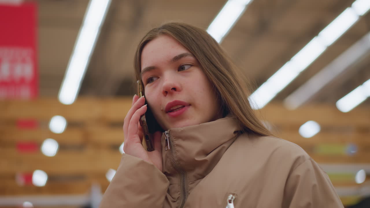 갈색 재을 입은 소녀의 클로즈업, 쇼핑몰에 서서 귀에 전화를 들고, 그녀의 얼굴에는 약간 불행한 표정이 있습니다.