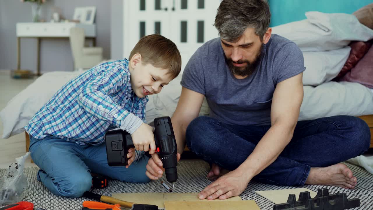 el niño está aprendiendo a usar un destornillador eléctrico mientras su padre le explica cómo trabajar con una pistola de tornillo y arreglar un tornillo en una pieza de madera.