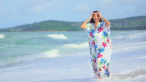 Cheerful-smiling-asian-woman-barefoot-walking-on-tropical-summer-beach-while-coming-waves-flooding-over-her-legs-on-a-sunny-day