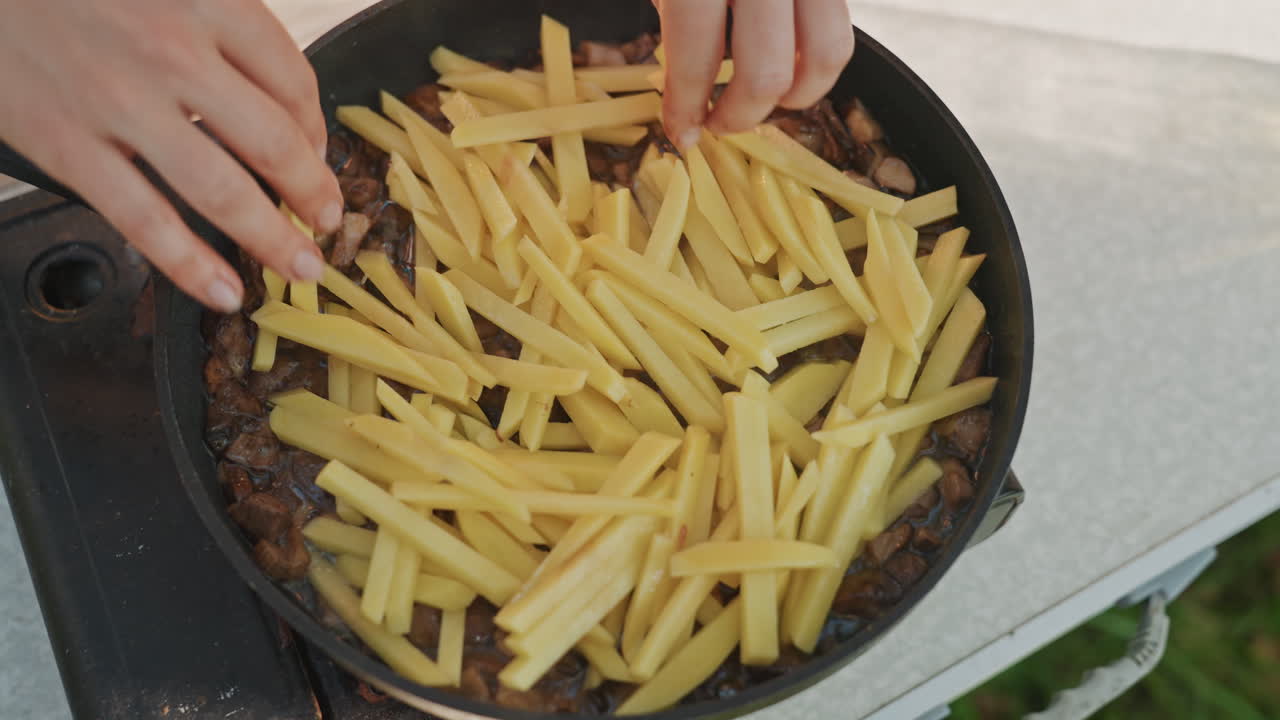 Hands placing fries over beef skillet on portable camping stove, outdoor picnic scene, sizzling meat browning under fries, tactile prep by camper, smoky aroma, rustic summer dinner shared at campsite