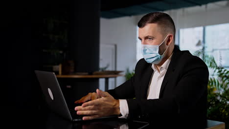 busy man working on his laptop. serious office worker wearing suit and mask typing on his computer. dark office backdrop.