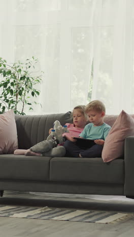 children boy and girl watch educational cartoons on tablet sitting on sofa against bright window. siblings use gadget preparing for school at home