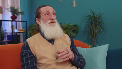 Thirsty-senior-old-man-sitting-at-home-holding-glass-of-natural-aqua-make-sips-drinking-still-water