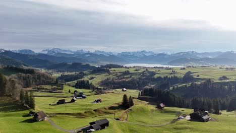 Flying-above-green-hilly-meadow-with-buildings-toward-lake-and-mountains