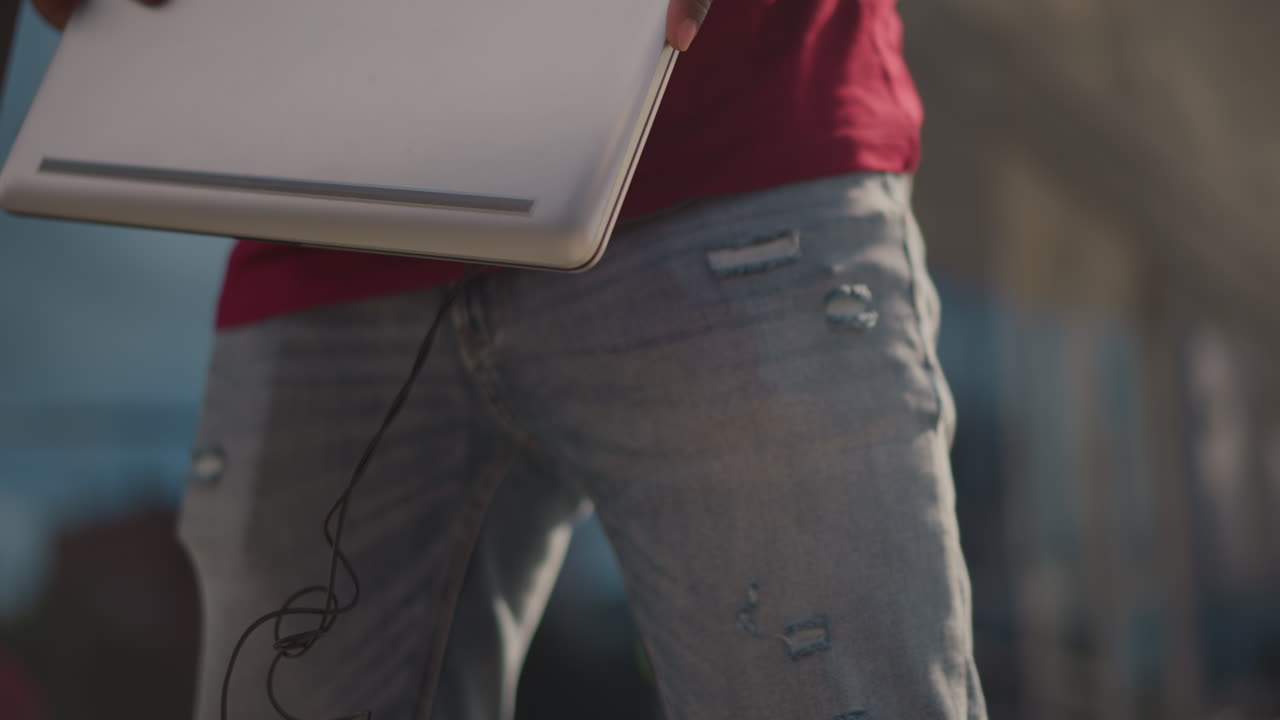 dj artist holding laptop and headphones steps across plaza, placing laptop into red sleeve, sliding controller case, adjusting headphones around neck, determined expression, shallow depth of field