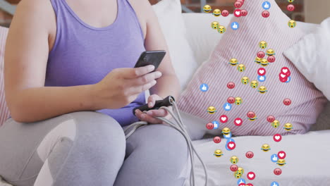 animation of emojis, like and love icons over woman using smartphone, during exercise at home