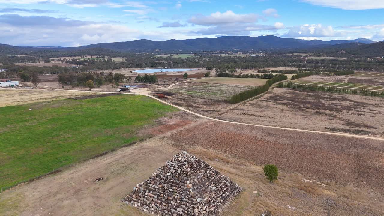 Drone camera smoothly flies over a stone pyramid structure in an open rural landscape near Ballandean, Queensland, under bright daylight with clear skies