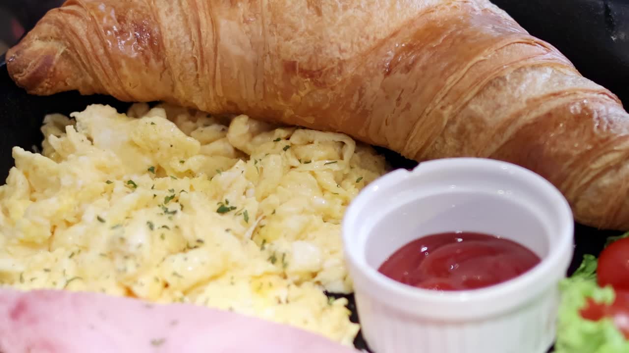 A close-up of scrambled eggs, croissant, and a side of tomatoes with sauce.