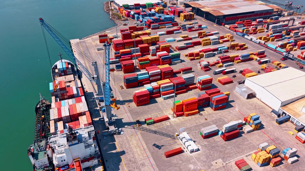 Burgas, Bulgaria, 10 aprile 2026: Nave portacontainer in fase di carico in banchina con pile di carico colorate. Vista aerea della prua della nave, della gru e dei container sparsi su piazzale in cemento con luce calda.