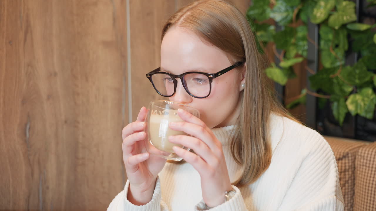 young lady holds glass of smoothie with both hands, eyes closed in quiet appreciation as she enjoys peaceful moment at wooden table with cake nearby in warm, cozy cafe environment
